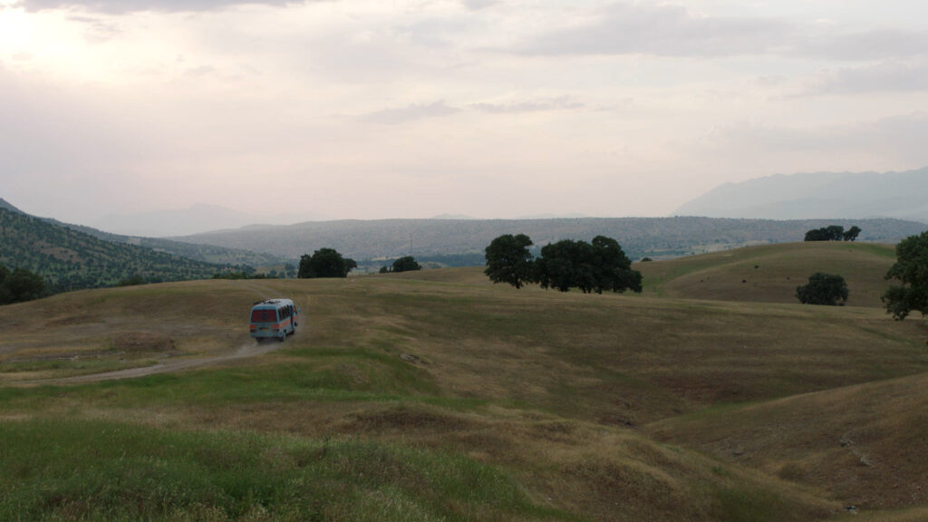 Hügelige Landschaft im zentraliranischen Plateau mit einem Mini-Van der eine Straße entlang in die Ferne fährt